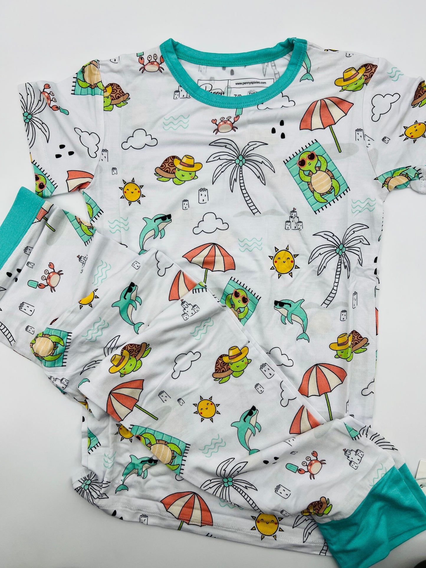 Flat lay of White bamboo kids pajamas featuring Tilly the Tortoise on the beach with Subtle Braille cues and a tag-free neckline support comfort and ease during everyday routines.