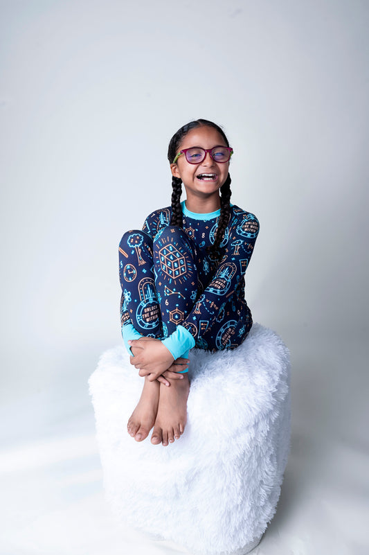 Young girl wearing a blue Math patterned Bamboo Pajamas and glasses, sitting on a white cushion against a light gray background.