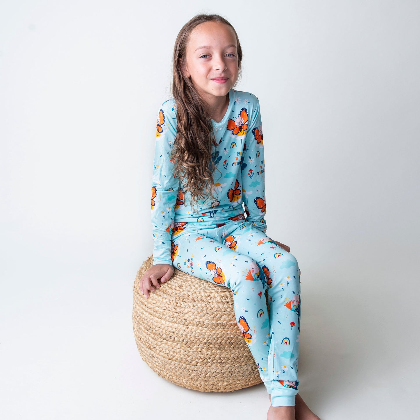 Young girl wearing a butterfly patterned pajama set sitting on a woven stool against a white background