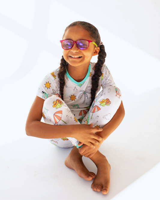 Girl sitting in a White bamboo kids pajamas featuring Tilly the Tortoise on the beach with Subtle Braille cues and a tag-free neckline support comfort and ease during everyday routines.