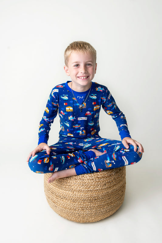 Young boy wearing blue pajamas with vehicle pattern sitting on a woven stool against a white background
