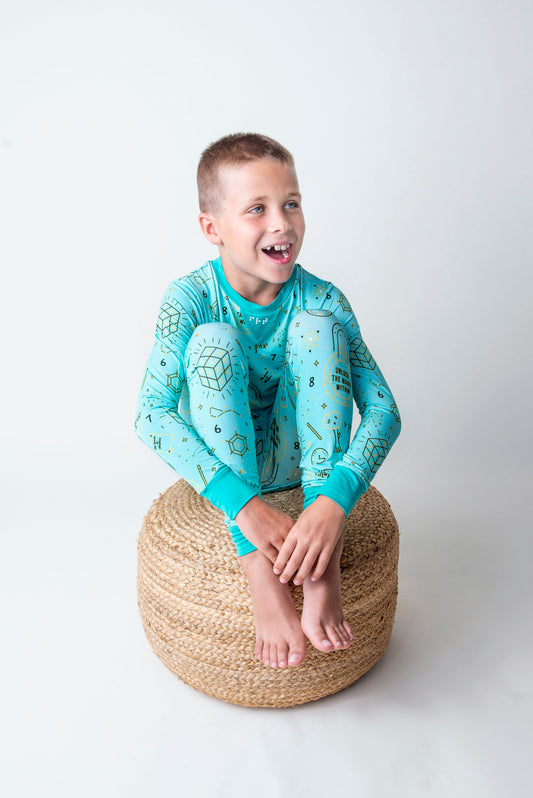 Child wearing a teal long-sleeve pajamas with geometric math patterns sitting on a woven stool.