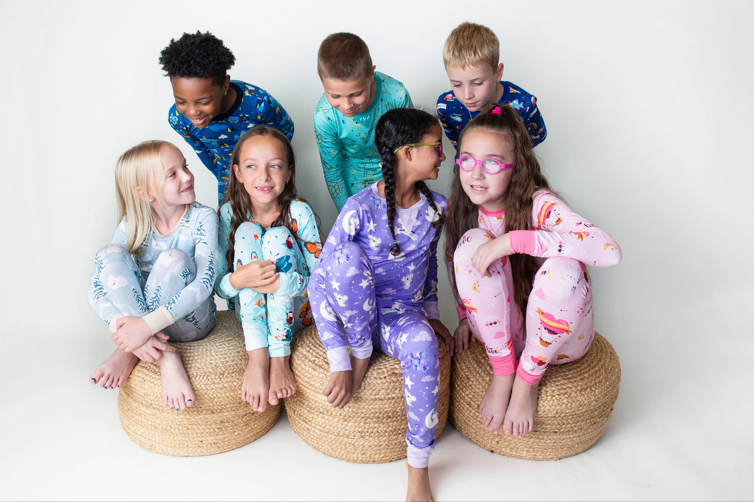 Children wearing colorful pajamas sitting on woven stools against a white background